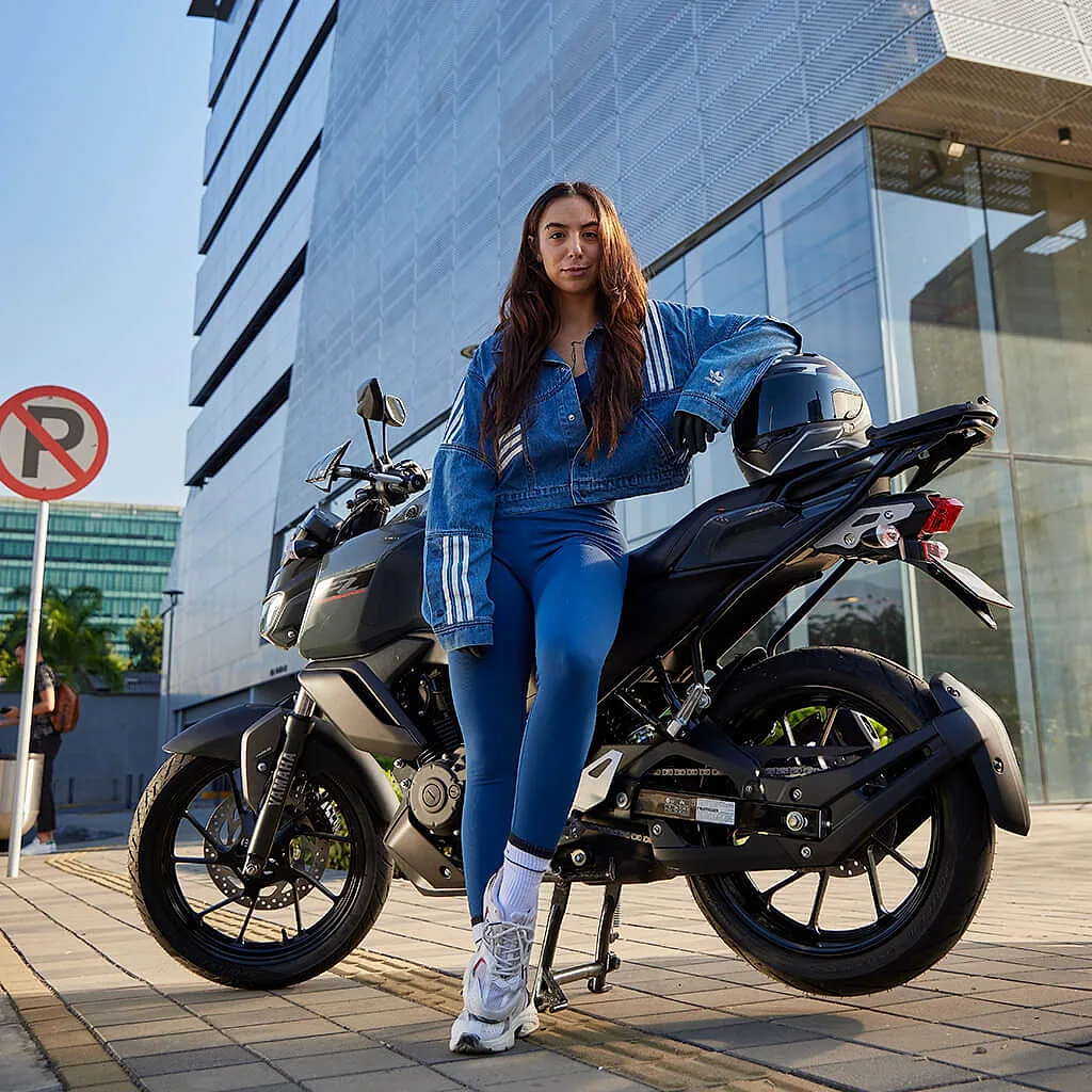 Mujer con chaqueta de mezclilla junto a una Yamaha FZ15 frente a un edificio de vidrio